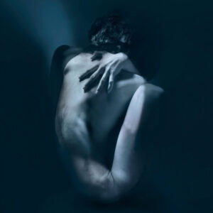 Fine art self-portrait in deep blue tones. A nude male figure sits curled inward, viewed from behind, with his head bowed and one hand resting across his opposite shoulder. Soft light illuminates the contours of his back and arms, while his face and much of his body disappear into shadow. The surrounding darkness and indistinct edges create an atmosphere of solitude, vulnerability, and quiet introspection.